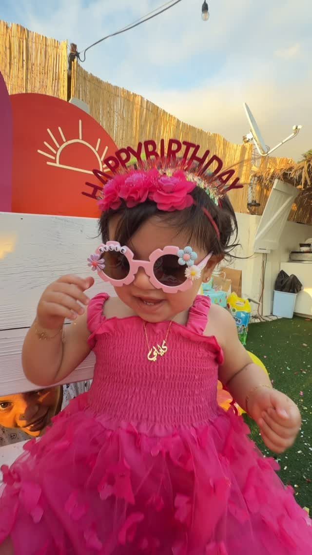 Baby girl in pink birthday dress and flower sunglasses posing outdoors for party.