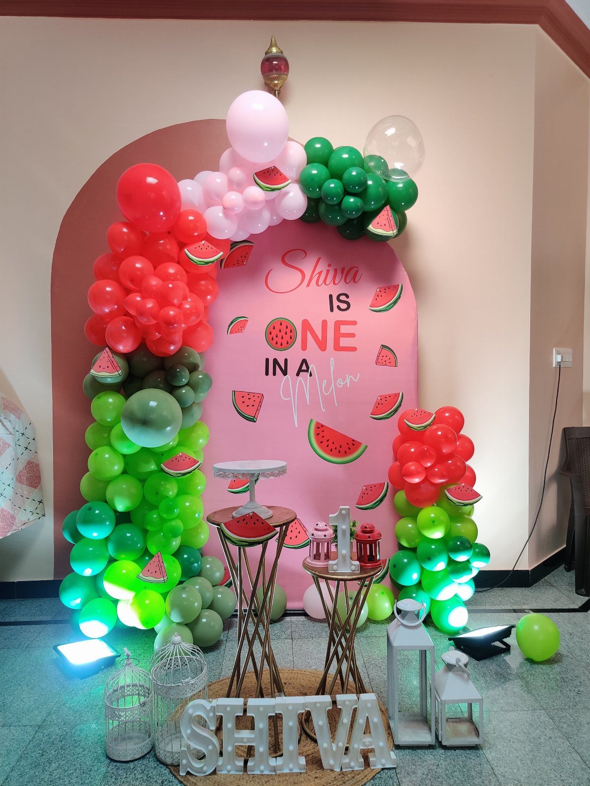 Watermelon themed birthday backdrop with red and green balloons and fruit cutouts.