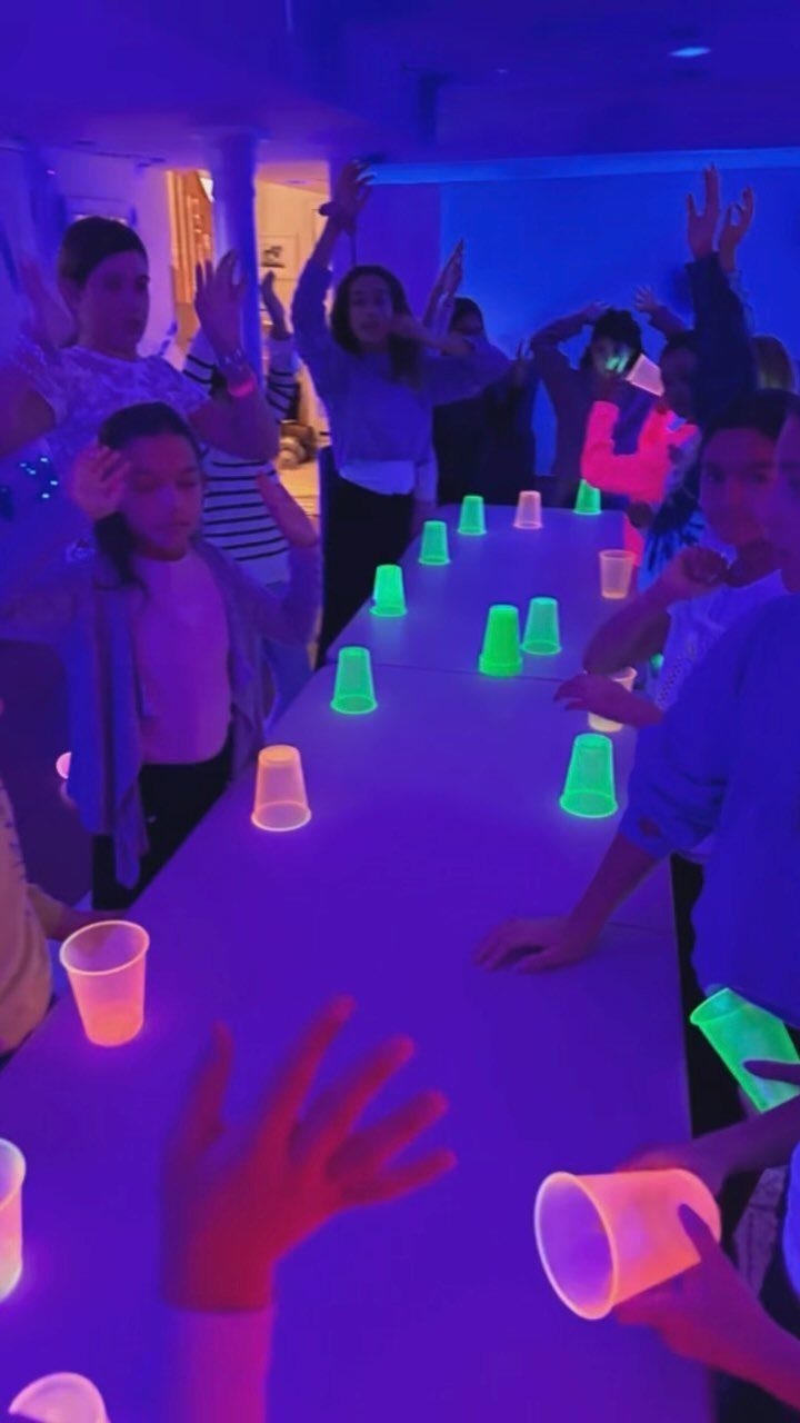 Young people partying around a table with glowing neon cups under UV light.