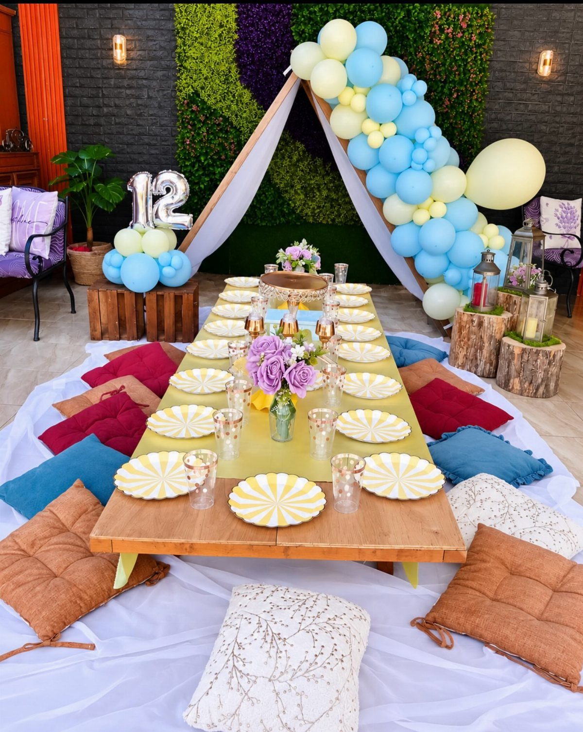 Low wooden picnic table with yellow plates, colorful cushions, and blue balloon arch.