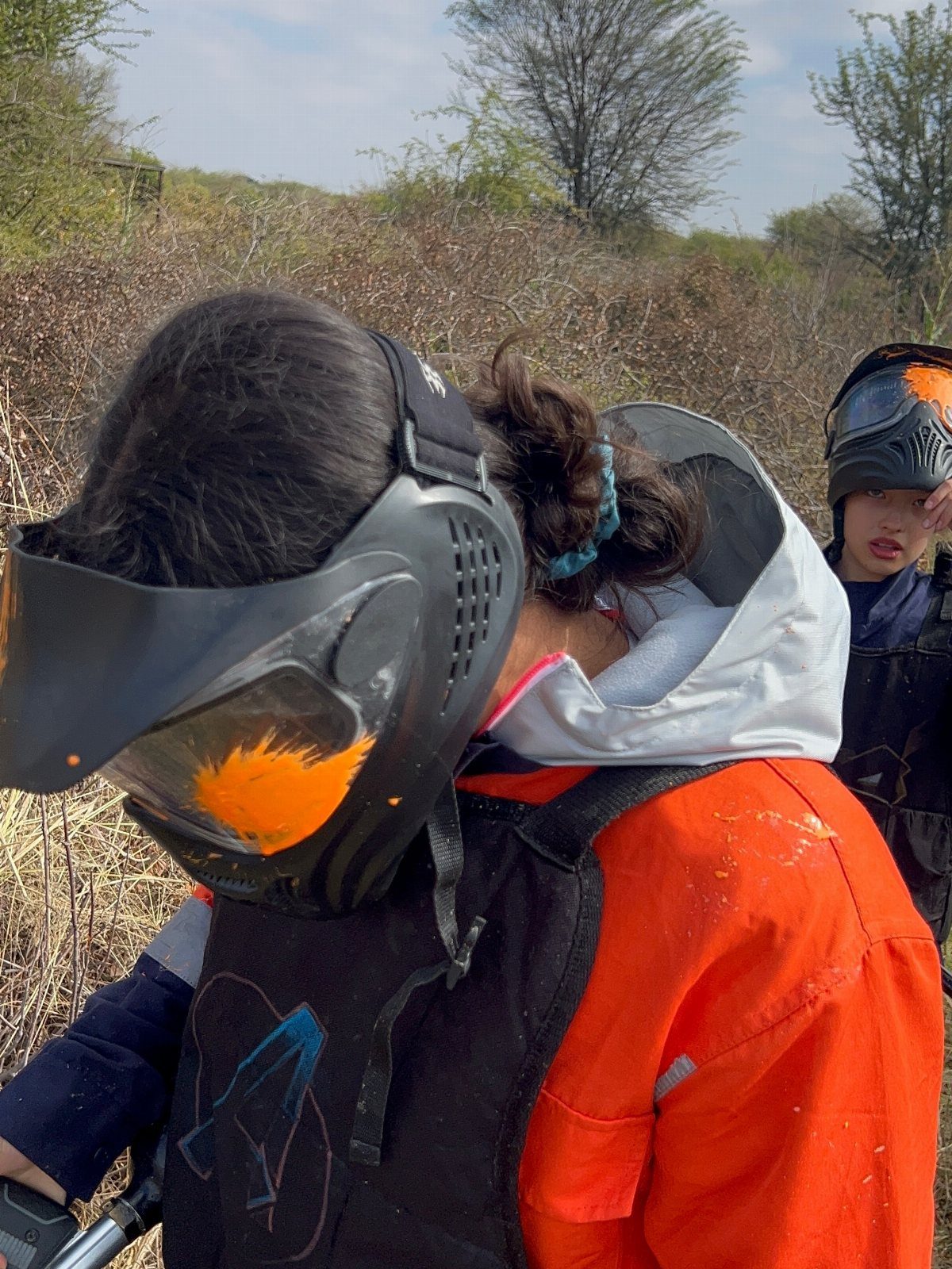 Children wearing paintball masks and protective gear in a dry outdoor field.