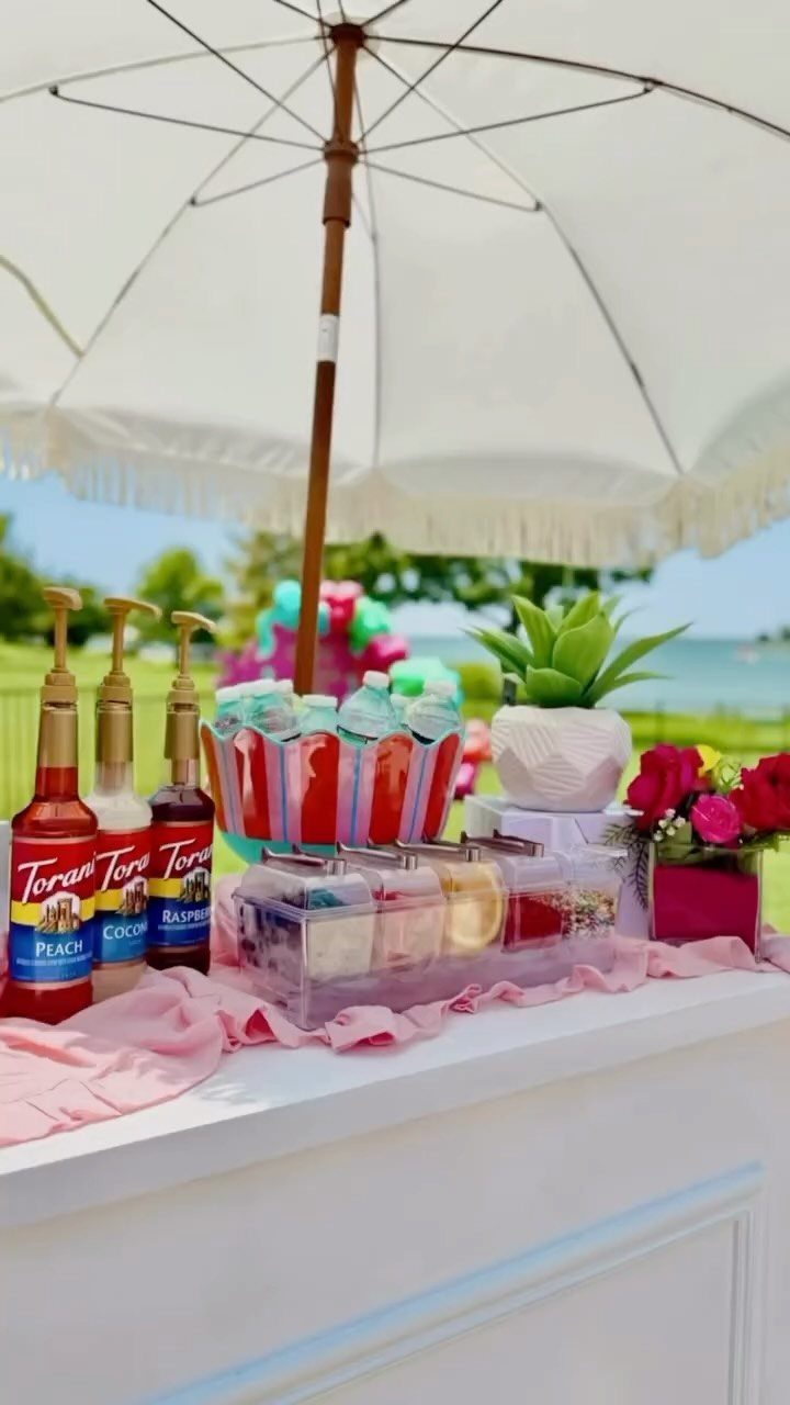 Outdoor beverage station with colorful drinks and syrups under a white fringed umbrella.