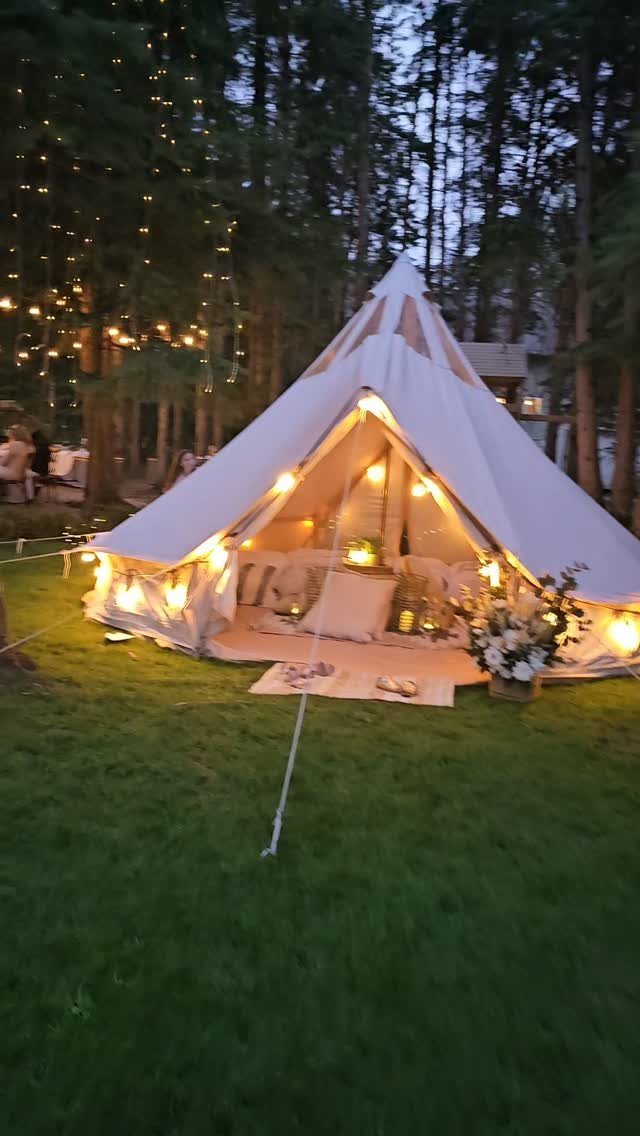White bell tent with warm interior lighting on a lawn surrounded by trees