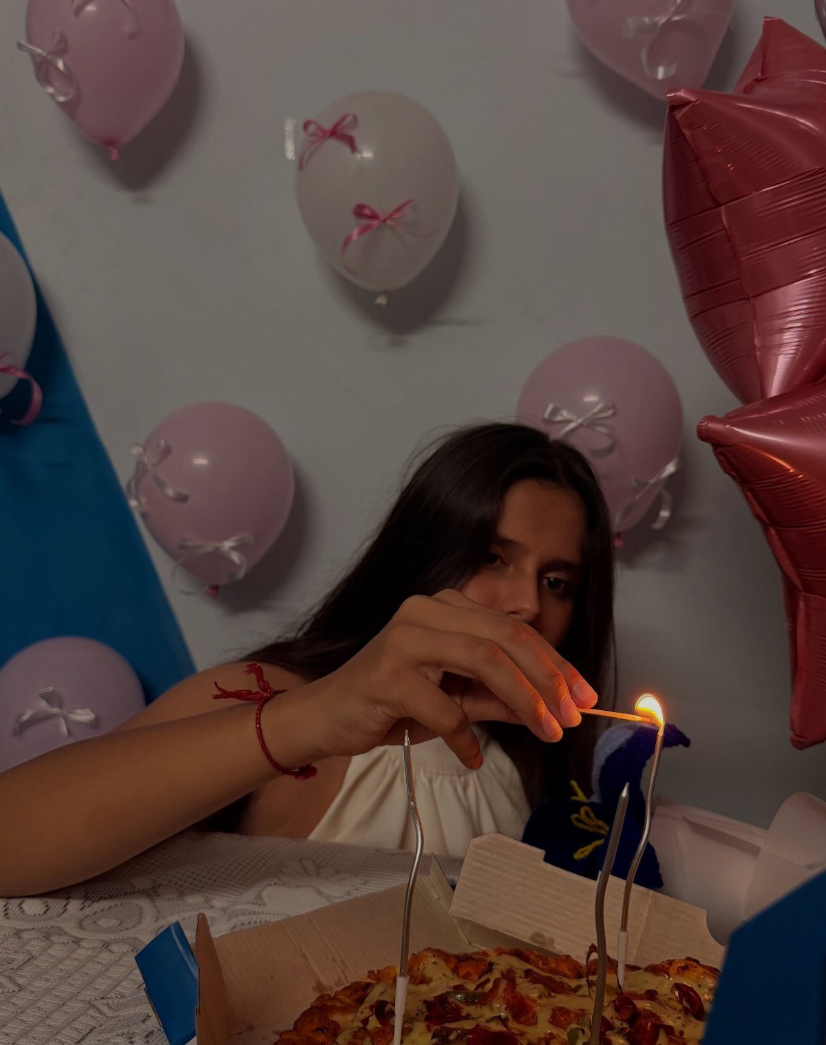 Teenage girl lighting candles on a pizza surrounded by pink and white balloons.