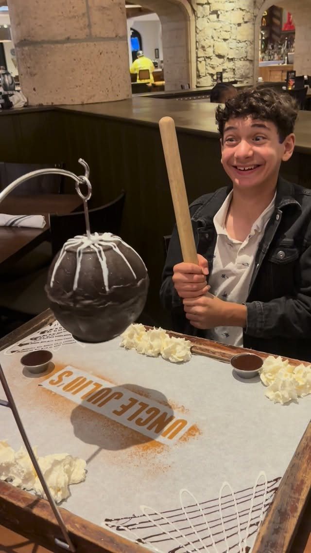 Teenage boy smiling while holding a wooden mallet over a chocolate dessert sphere.