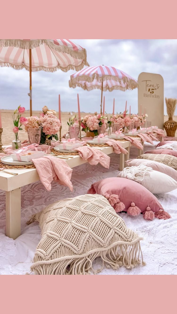 Beach party setup with a low table, pink decor, and striped umbrellas on sand.