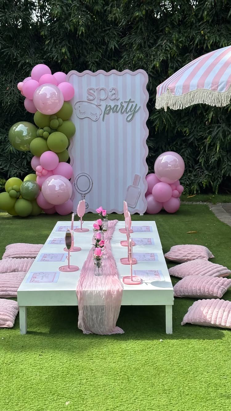 Outdoor spa party setup with a white table, pink cushions, and balloon arch.