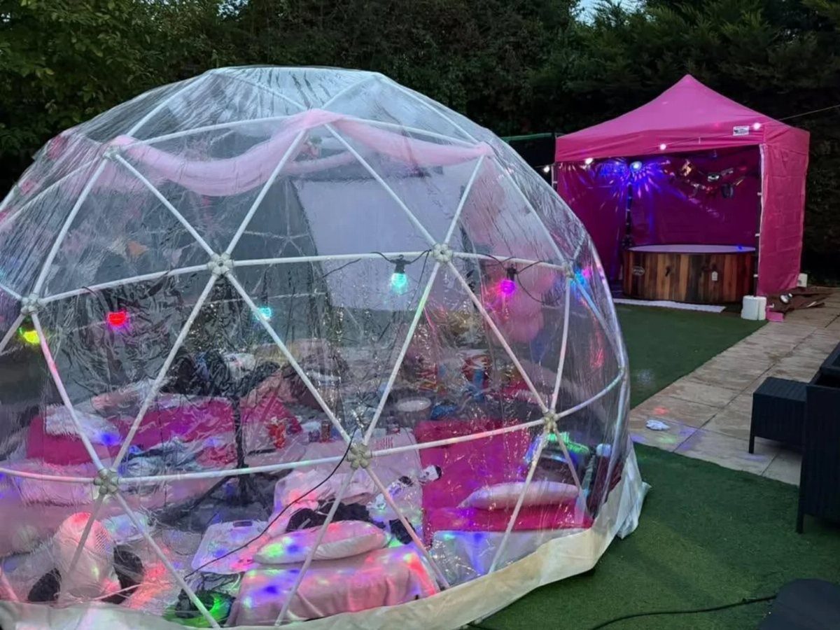 Clear geodesic dome tent with pink bedding and colorful lights on a lawn