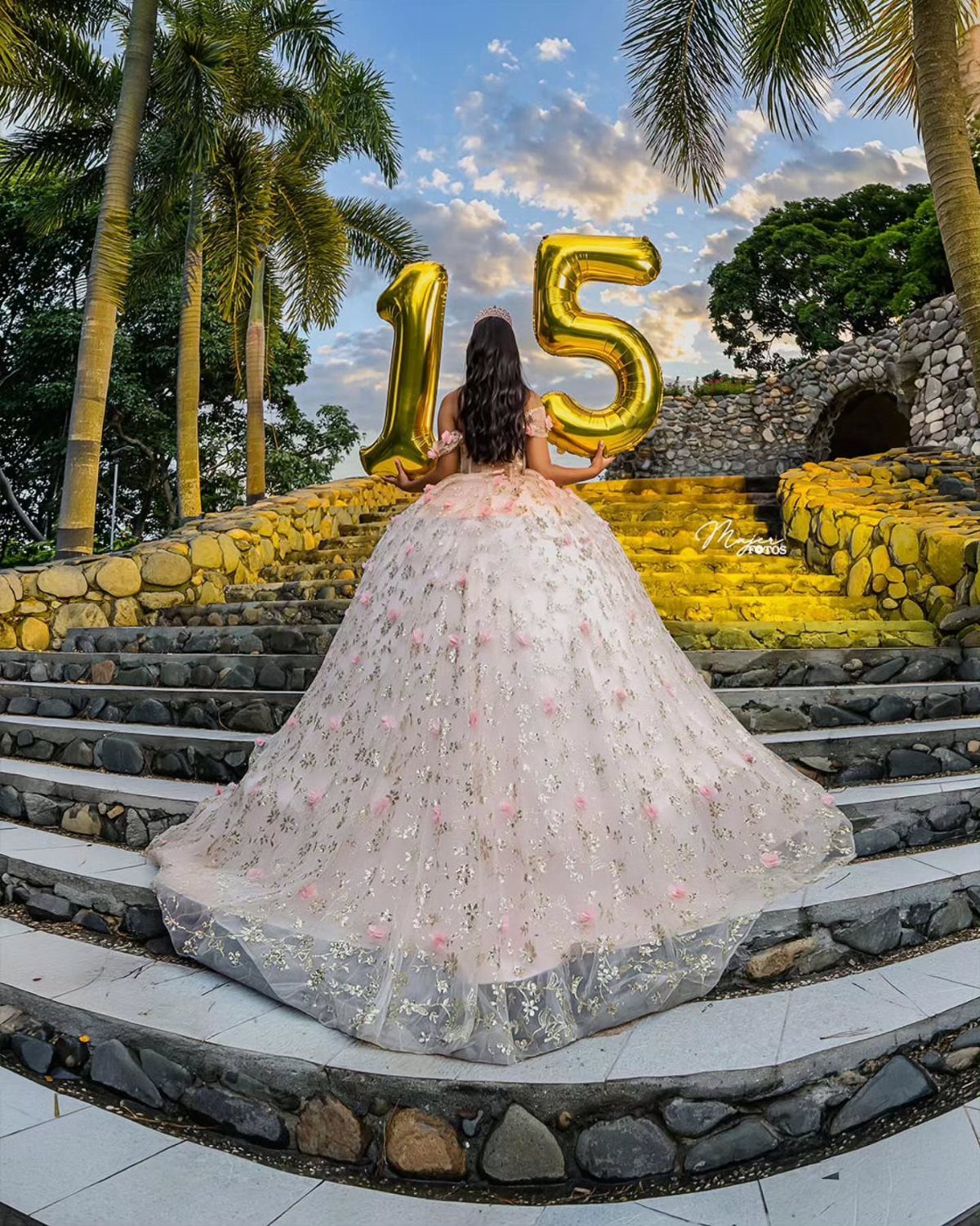 Girl in pink floral ballgown holding gold 15 balloons on stone steps.
