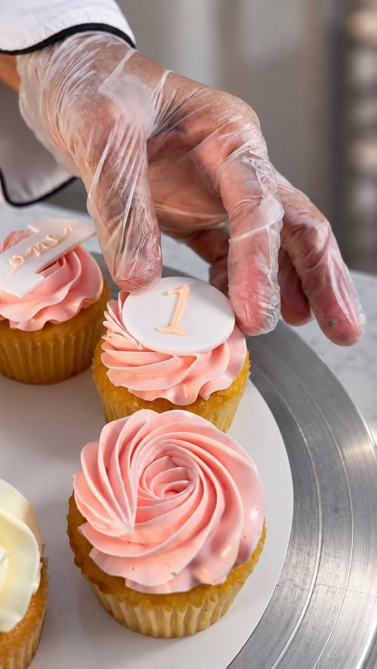 Cupcakes with pink swirled frosting and a number one topper being decorated.