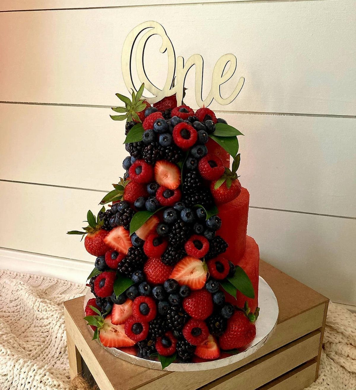 Fruit-covered red birthday cake with a wooden One topper on a wooden crate
