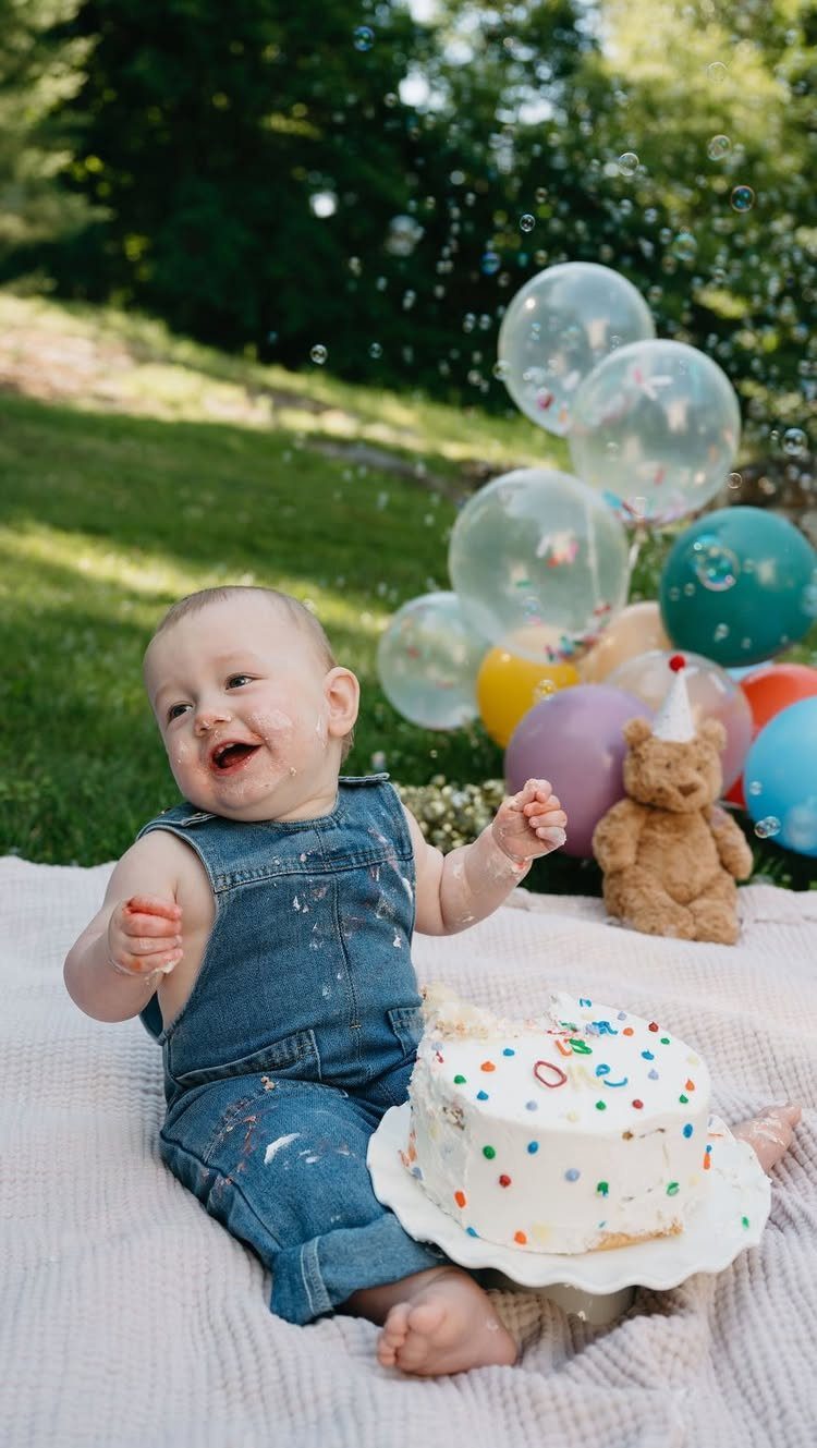 Baby in denim overalls smiling next to a small white sprinkle cake outdoors.