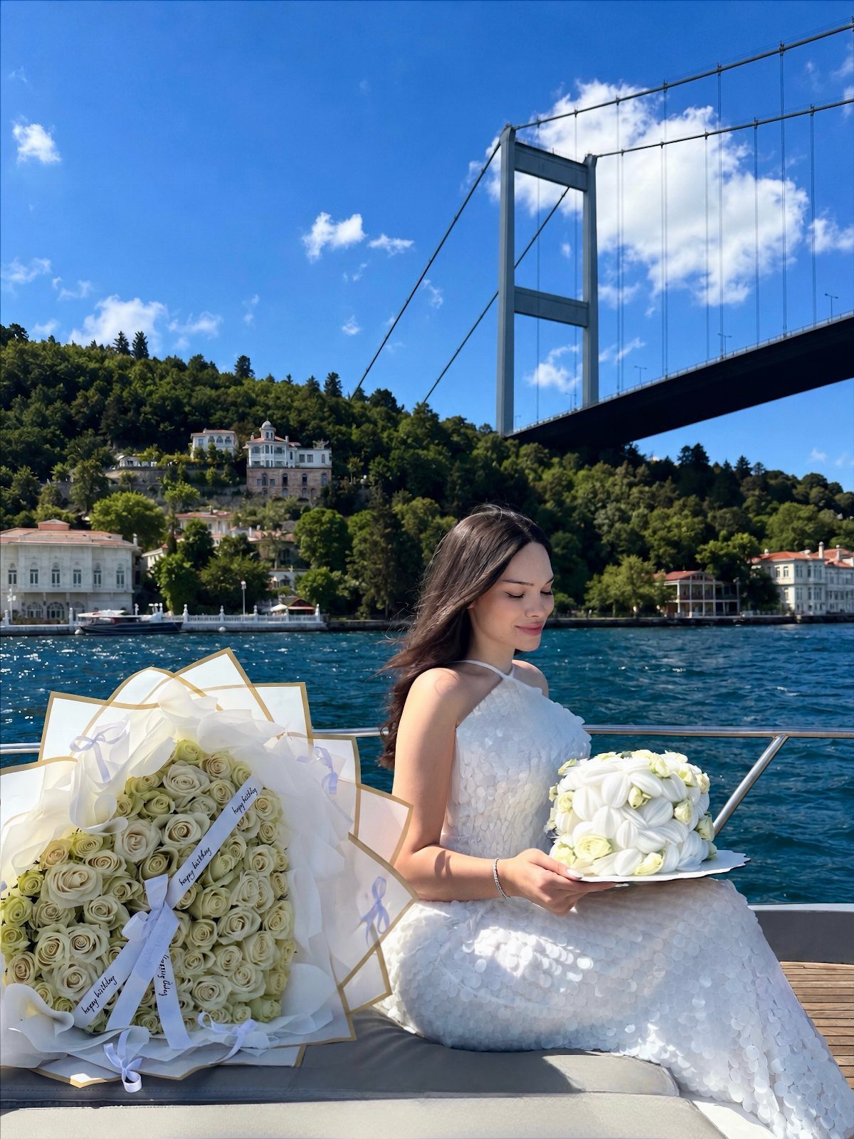 Woman in white dress holding a cake on a boat near a bridge.