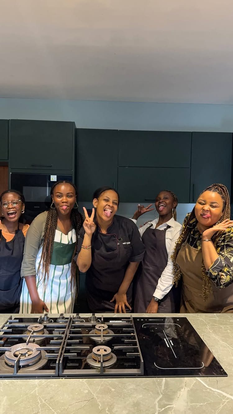 Five women in aprons posing and smiling in a modern kitchen setting.