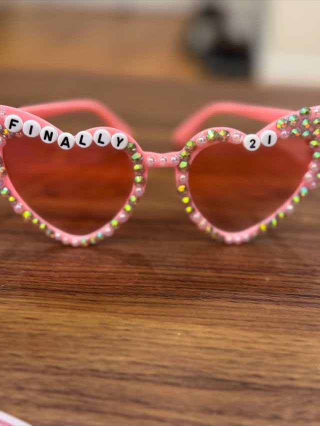 Pink heart-shaped sunglasses with rhinestones and beads spelling FINALLY 21 on wood.