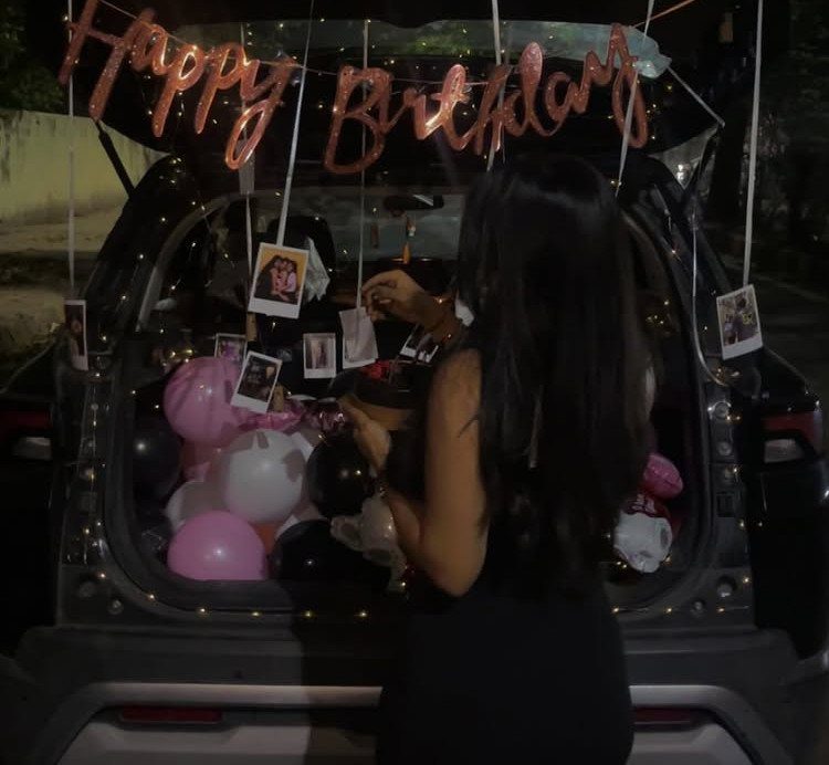 Woman standing by a car trunk decorated with balloons and birthday banners.