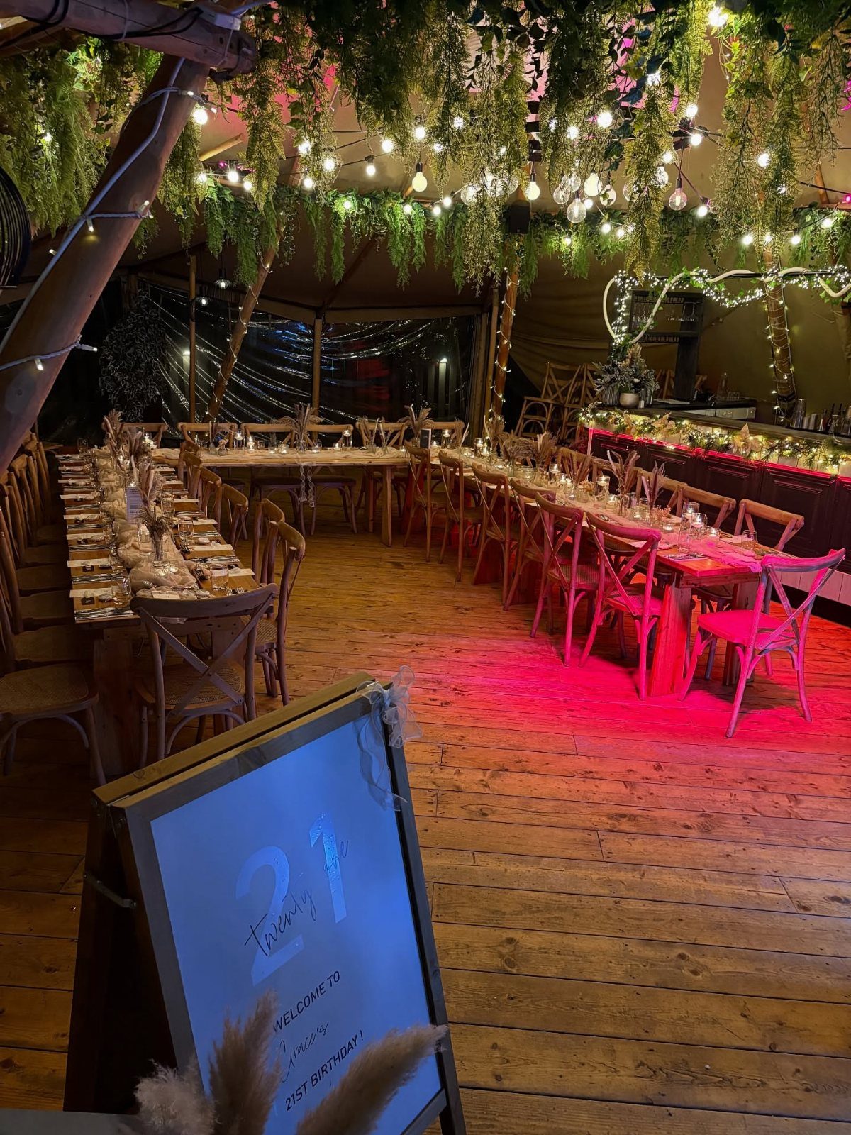 Long wooden dining table set for a 21st birthday party under hanging greenery.
