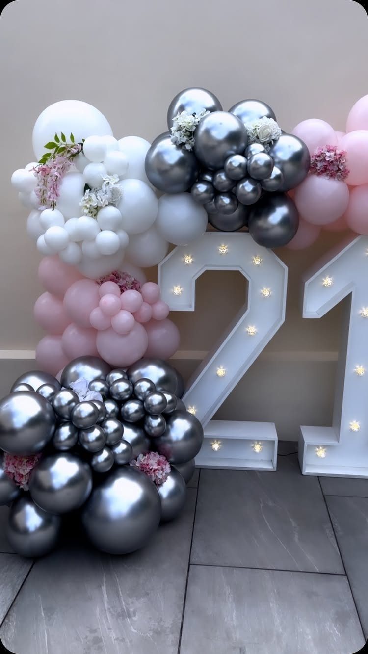 Large illuminated number 21 surrounded by silver, white, and pink balloon clusters.