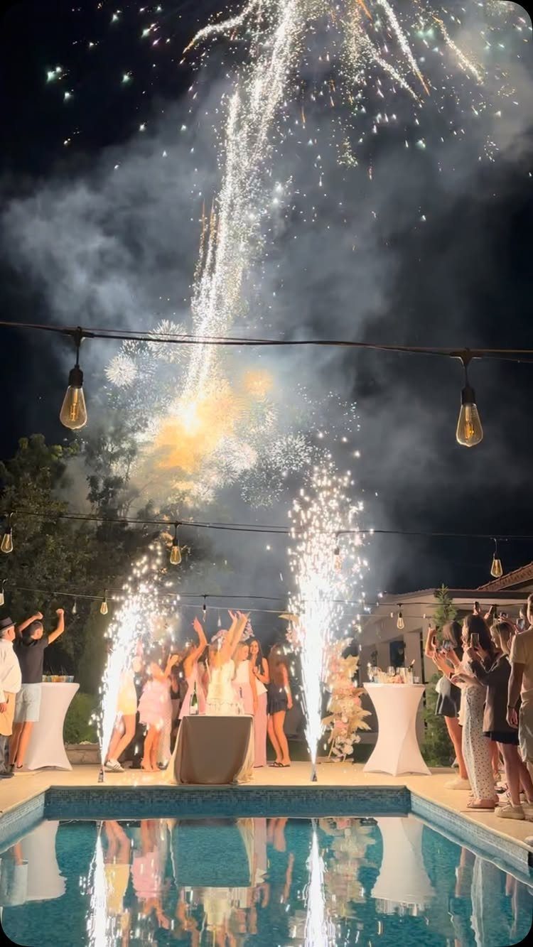 Nighttime pool party with fireworks and pyrotechnic sparks surrounding celebrating guests.