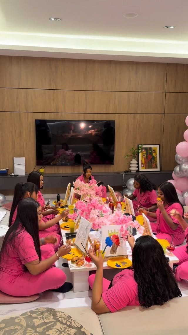 Women in pink pajamas painting on canvases at a decorated birthday party table.