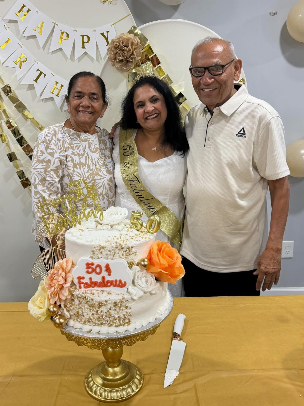 Three adults smiling behind a gold and white 50th birthday cake and decorations.