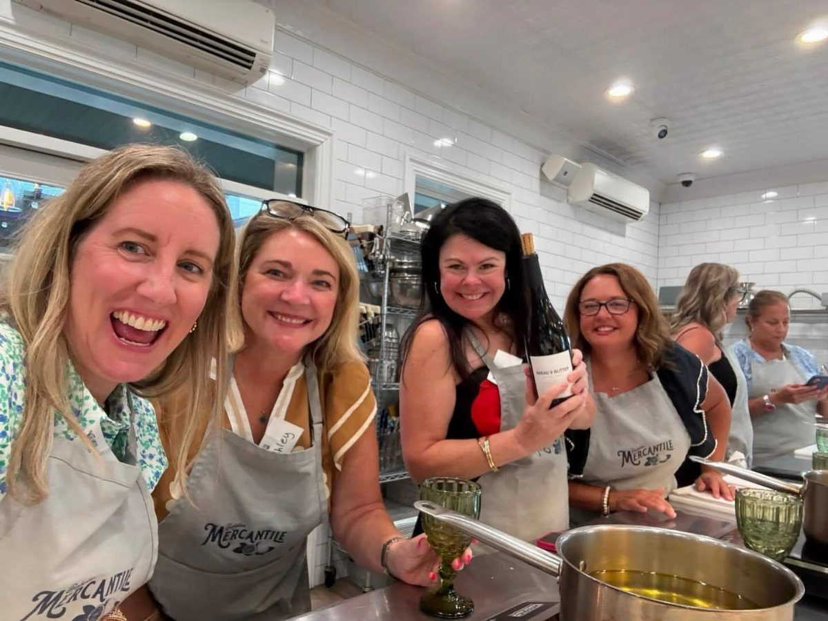 Four women in aprons smiling in a kitchen during a cooking class.