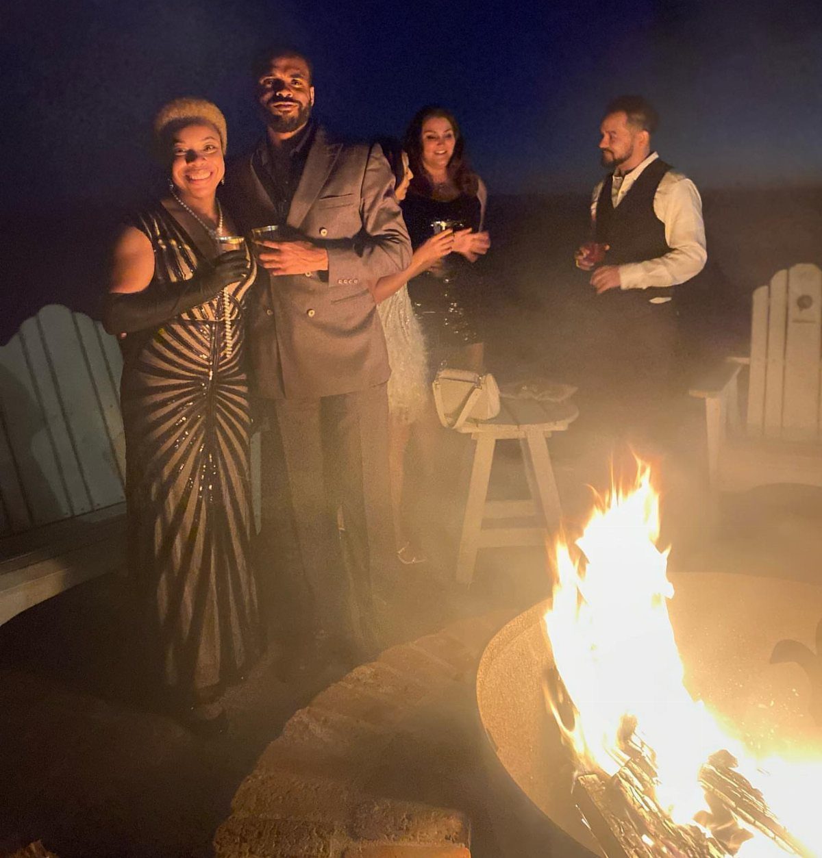 Formaly dressed adults standing and socializing around a glowing outdoor fire pit at night.