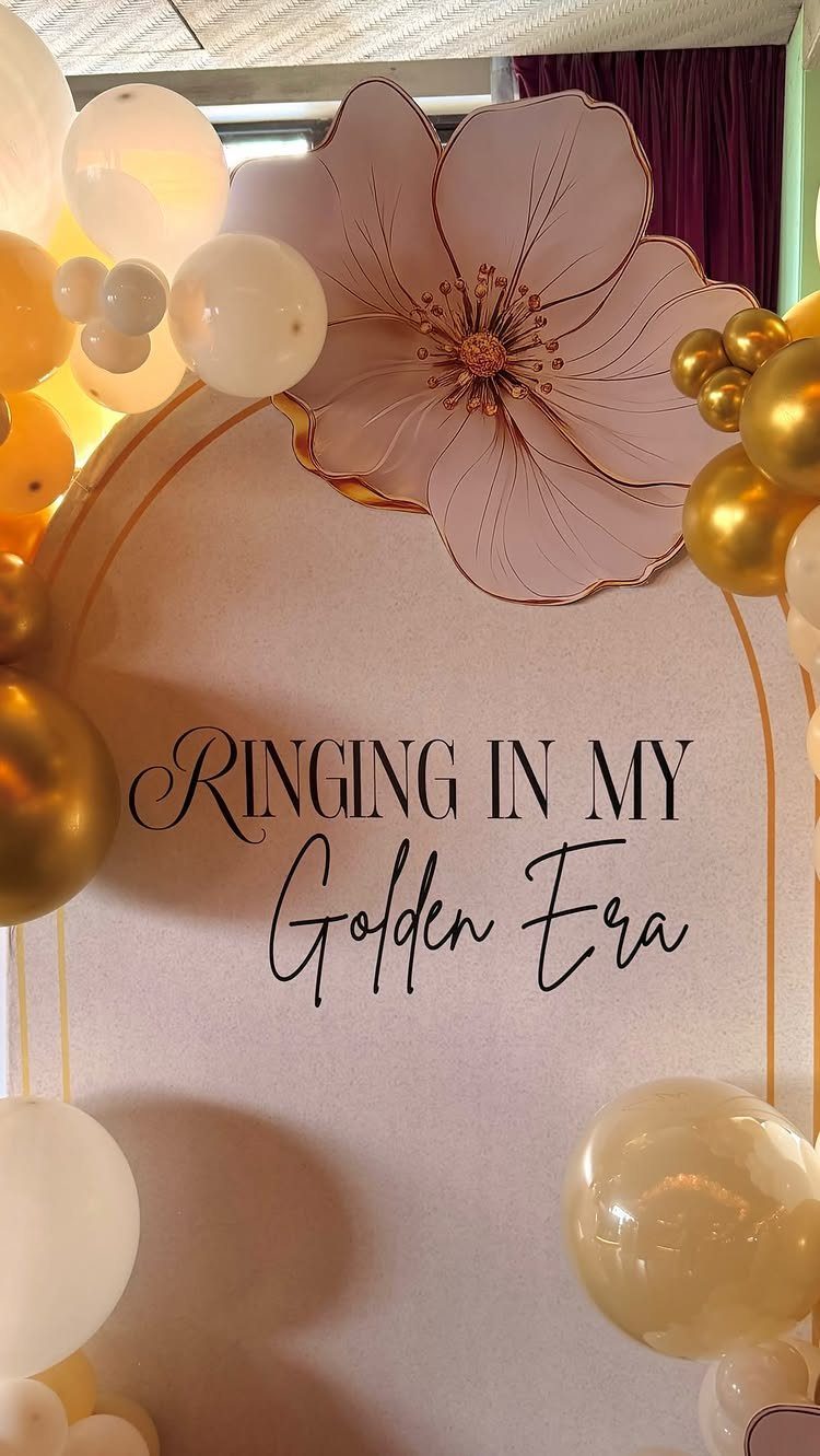 Party backdrop with gold and white balloons, a pink flower, and golden era text.