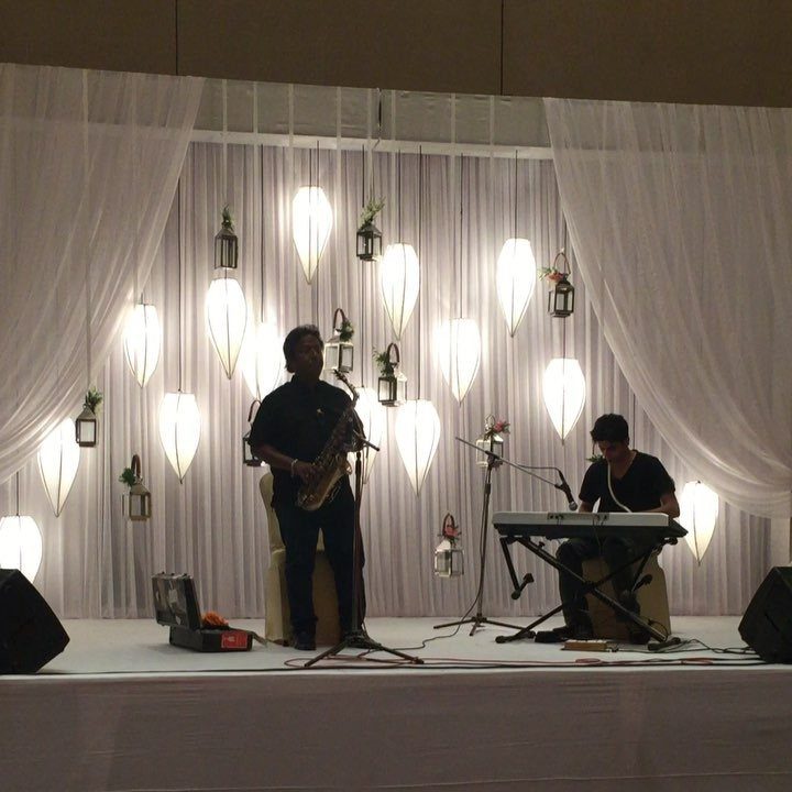 Two musicians playing saxophone and keyboard on a stage with hanging white lanterns.