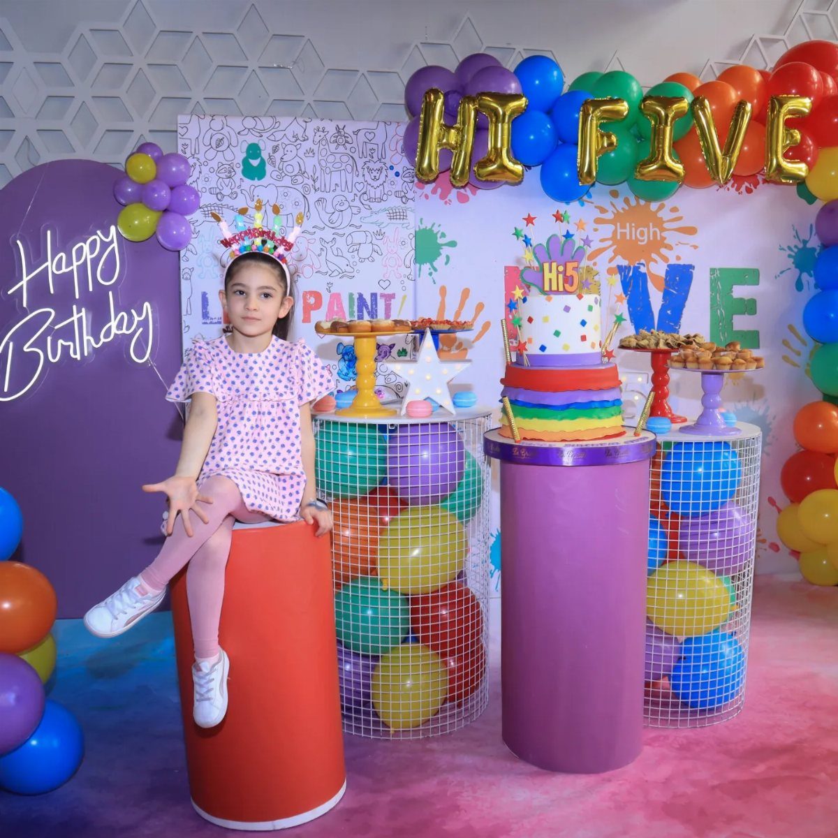 Young girl in a crown sitting amidst colorful balloons and a rainbow cake.