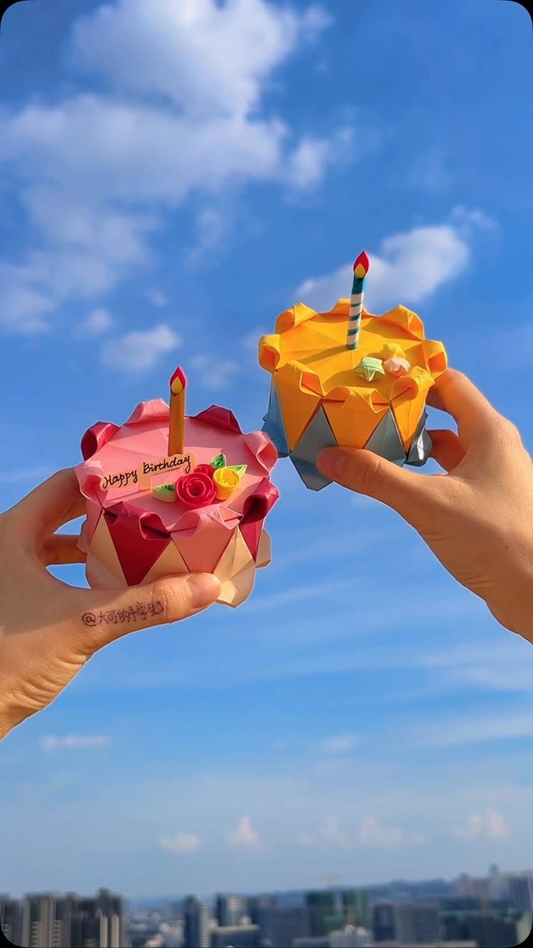 Two colorful origami paper cakes held up against a bright blue cloudy sky.