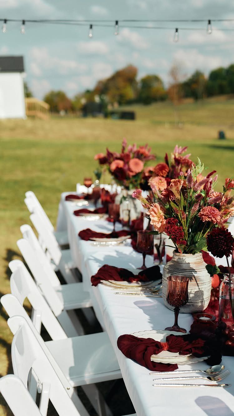 Long outdoor dining table set with autumn flowers and burgundy linens on green grass.