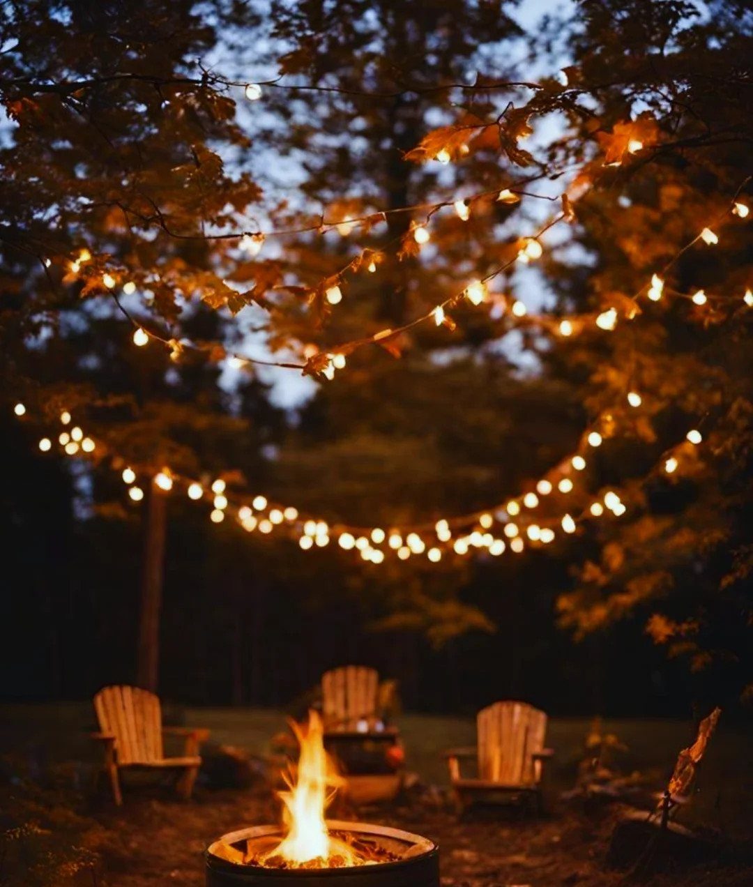 Fire pit with wooden chairs and string lights under autumn trees at night.
