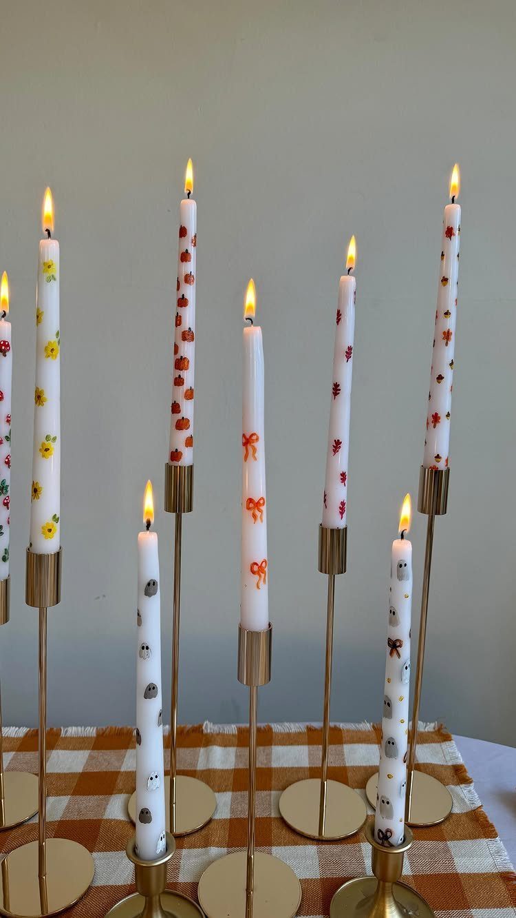Decorated white taper candles with autumn patterns in gold holders on checkered cloth.