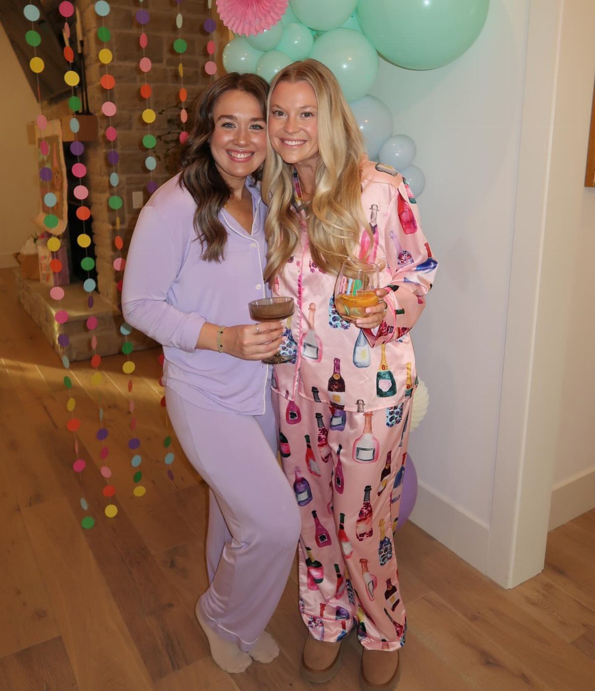 Two women in pajama sets holding cocktails in a decorated party room