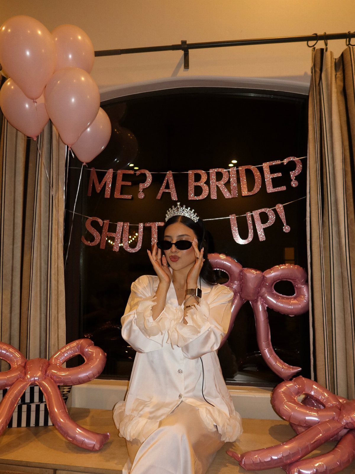 Woman in white satin pajamas and tiara posing with pink bridal party decorations