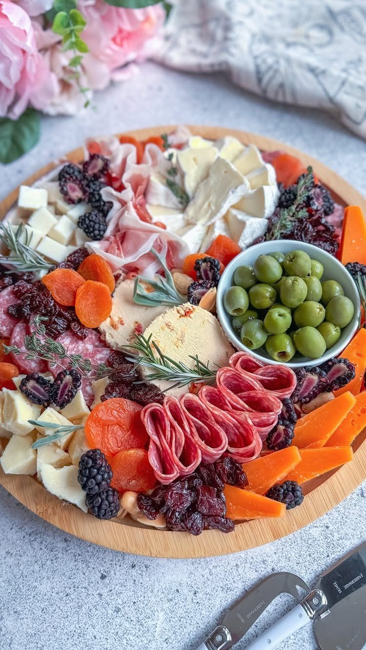 Wooden grazing board with assorted cheeses, meats, olives, and dried fruits on grey surface.
