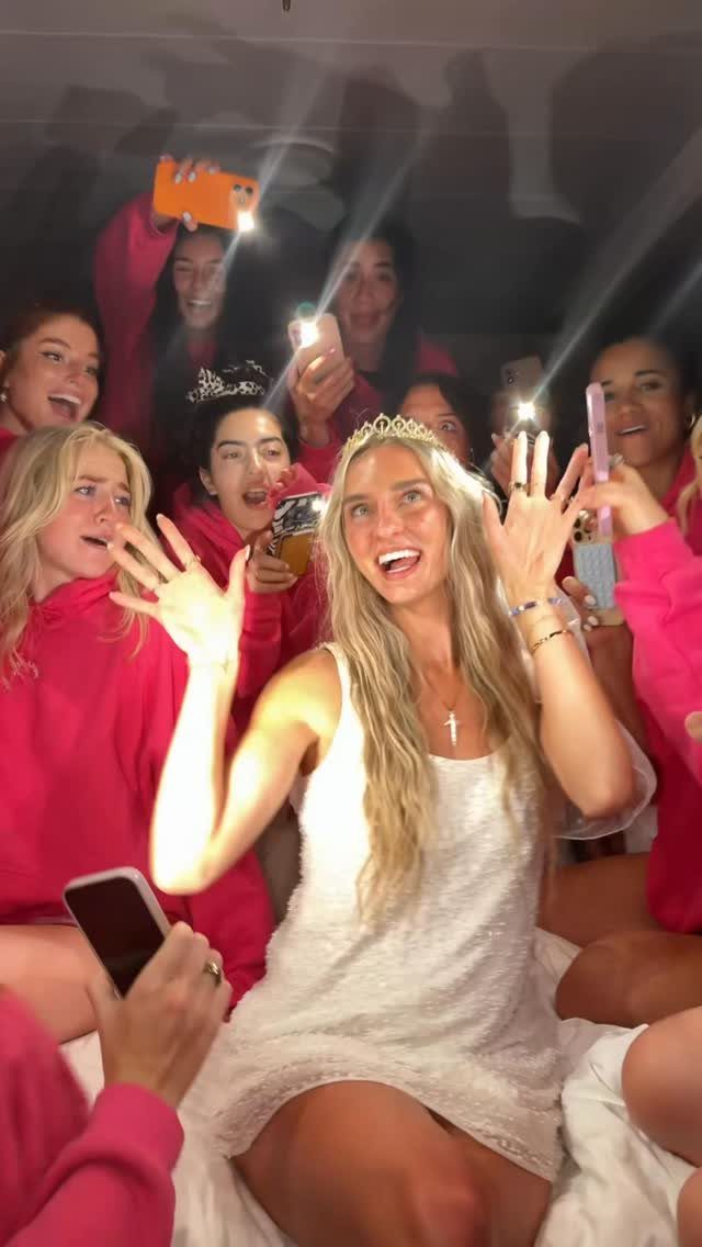 Bride in white lace dress and tiara surrounded by friends in pink hoodies.