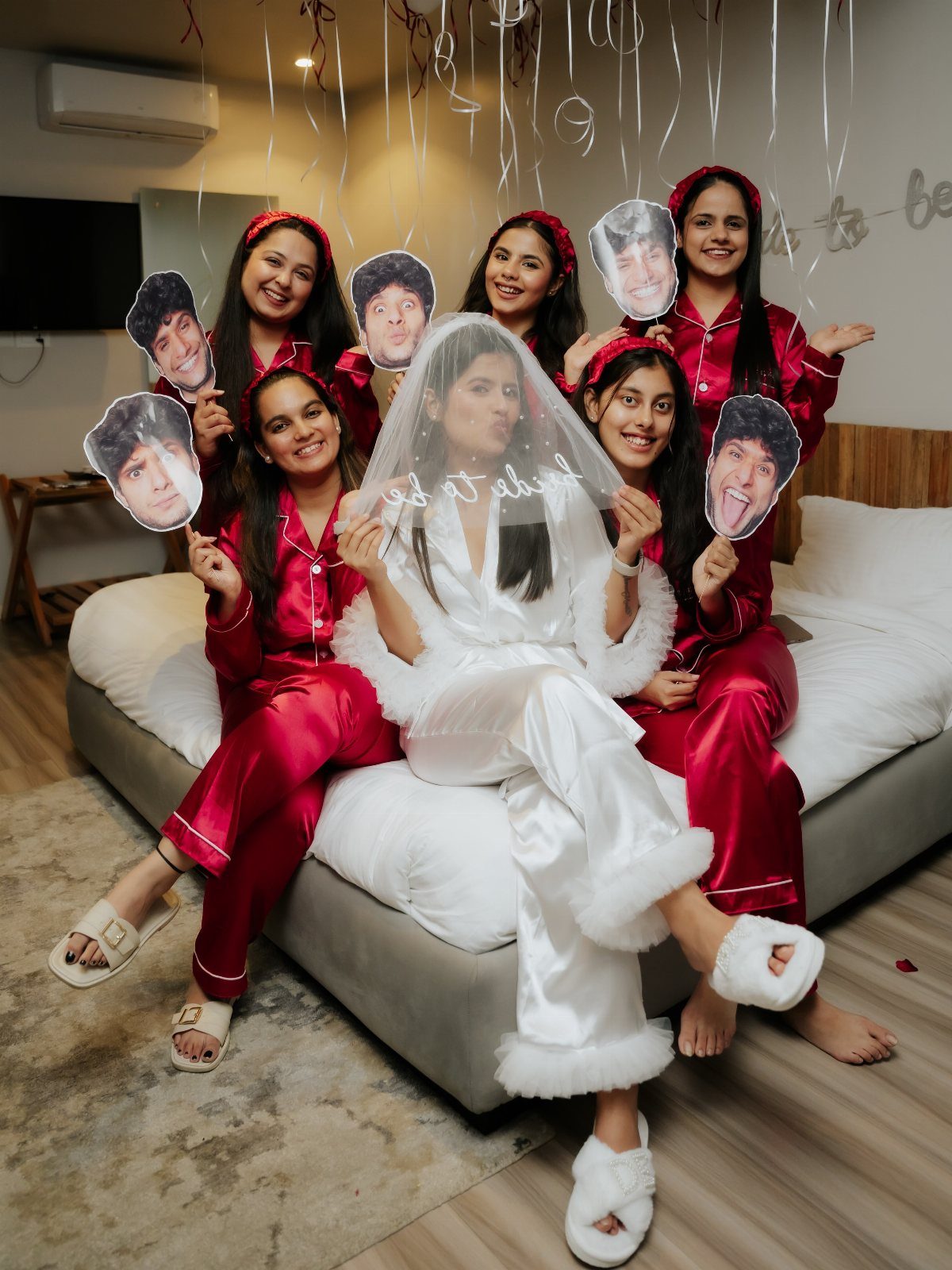 Women in red and white pajamas on a bed holding man's face cutouts.