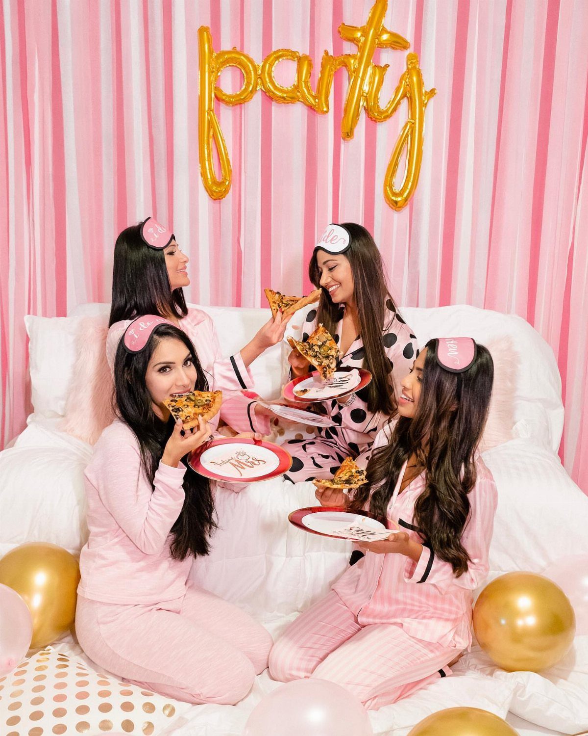 Four women in pajamas eating pizza in front of pink curtains and gold balloons.