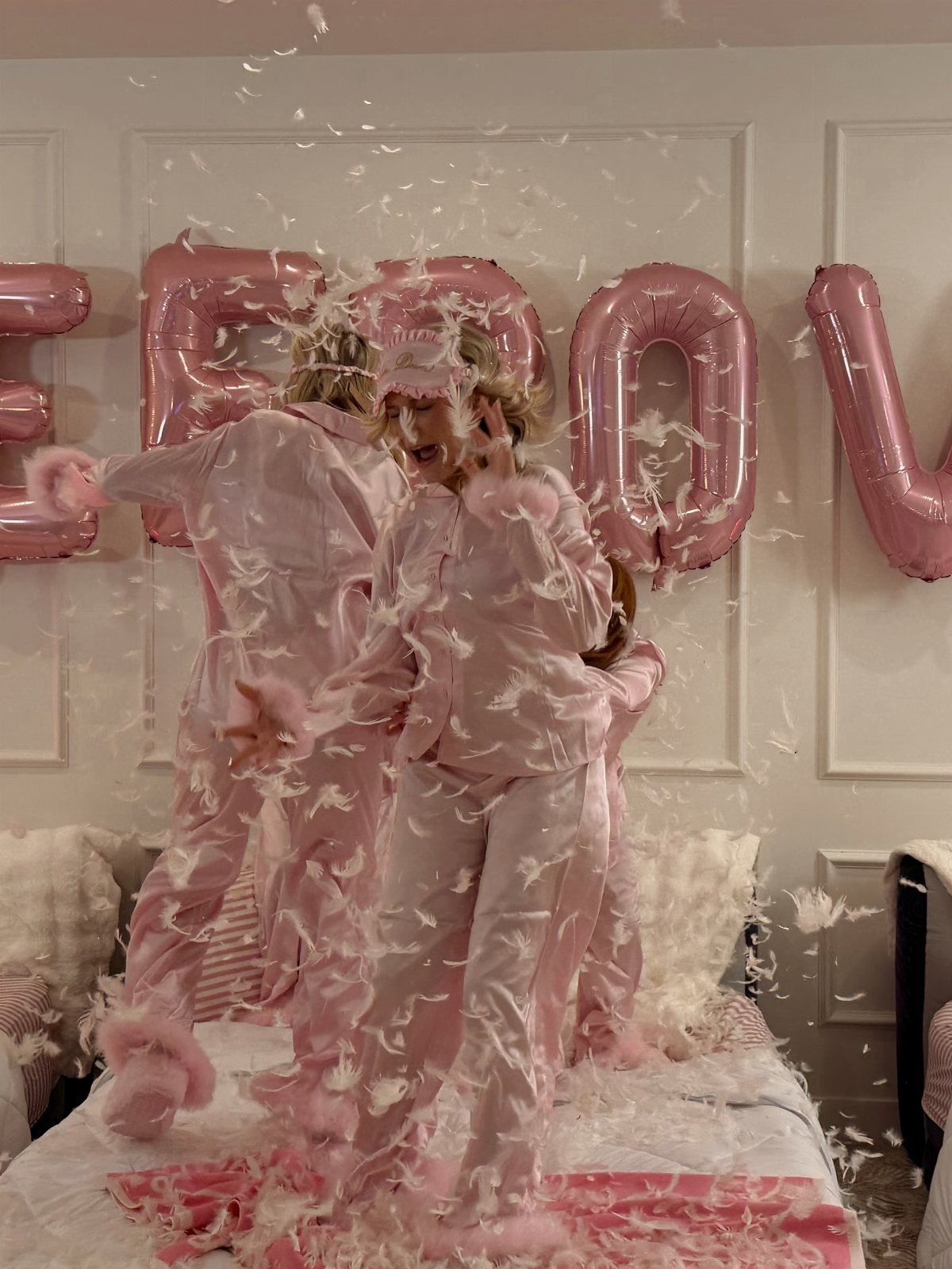 Two women in pink pajamas jumping on a bed with falling white feathers.