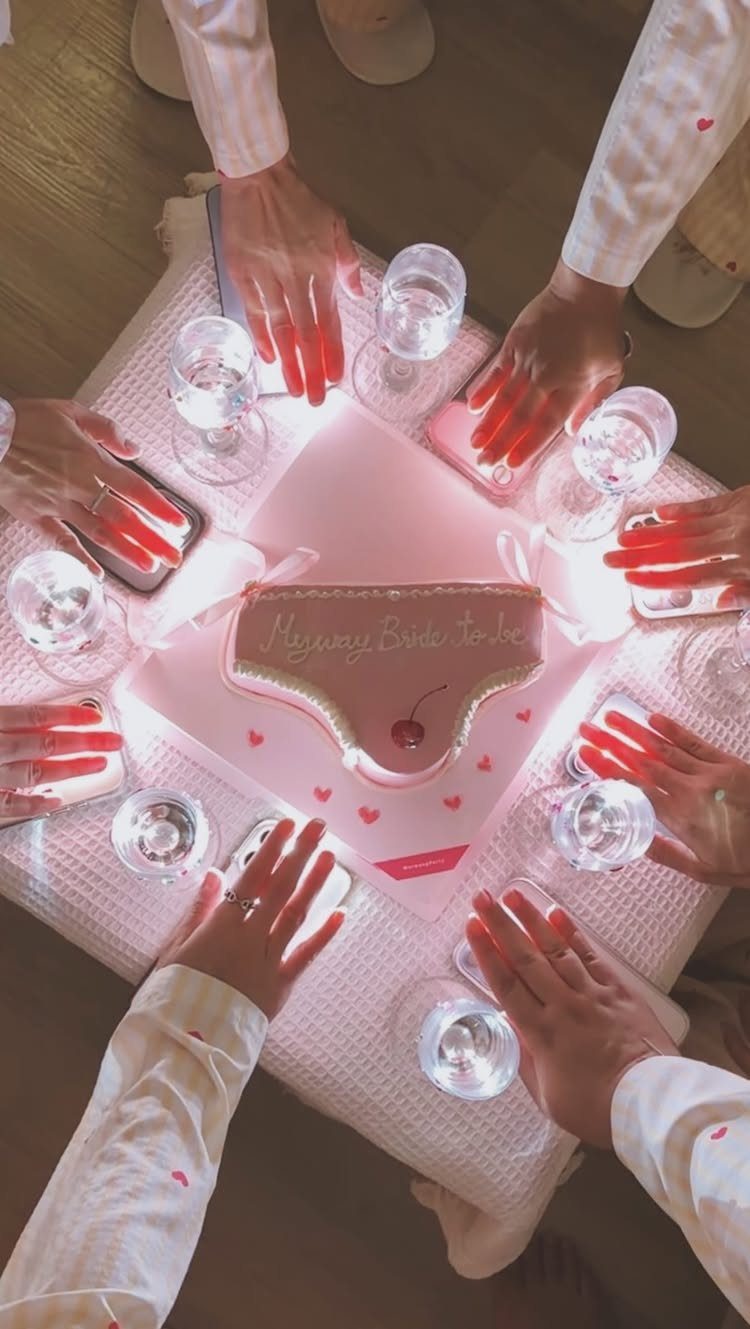 Women in white robes gathered around a pink panty-shaped bachelorette party cake.