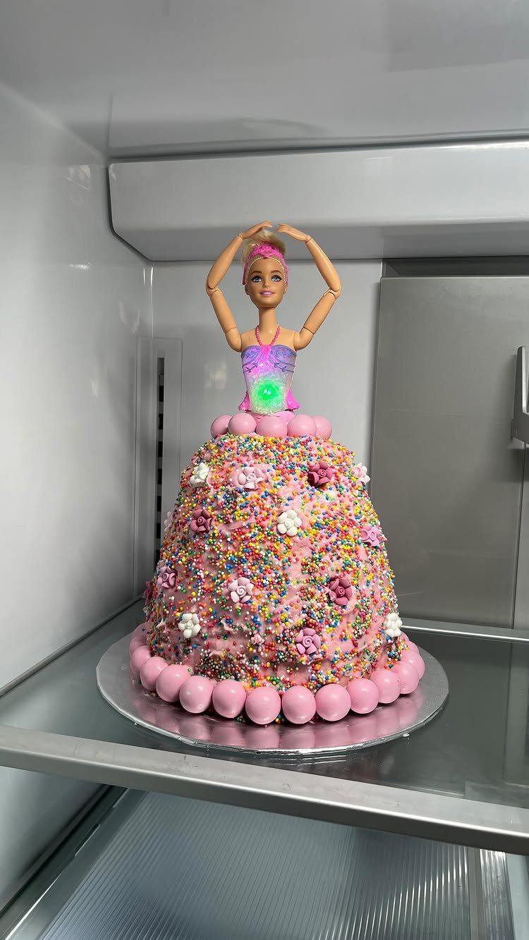 Barbie doll cake with pink frosting and colorful sprinkles inside a white refrigerator