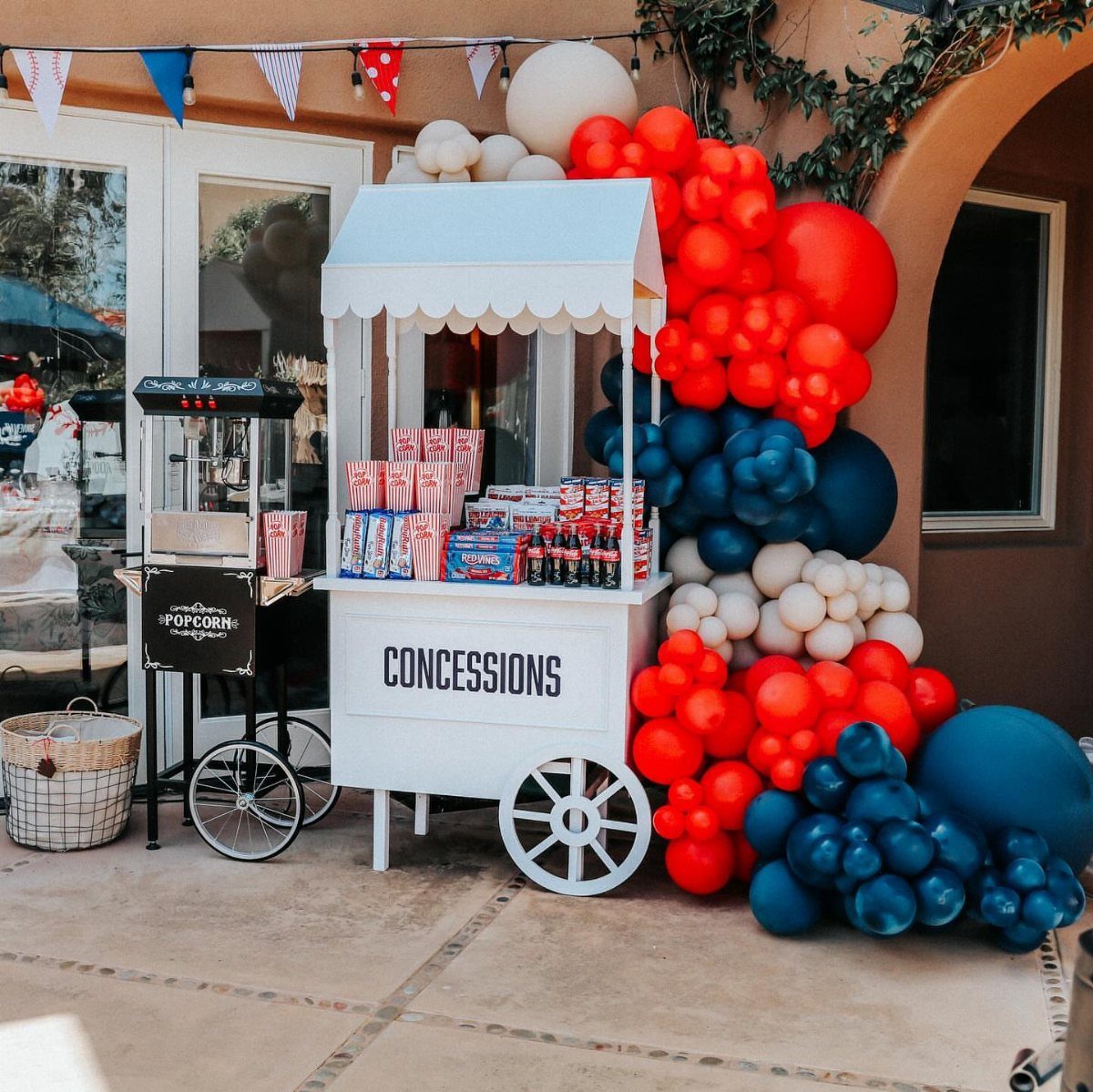 White concession cart with popcorn and drinks decorated with red and blue balloons.