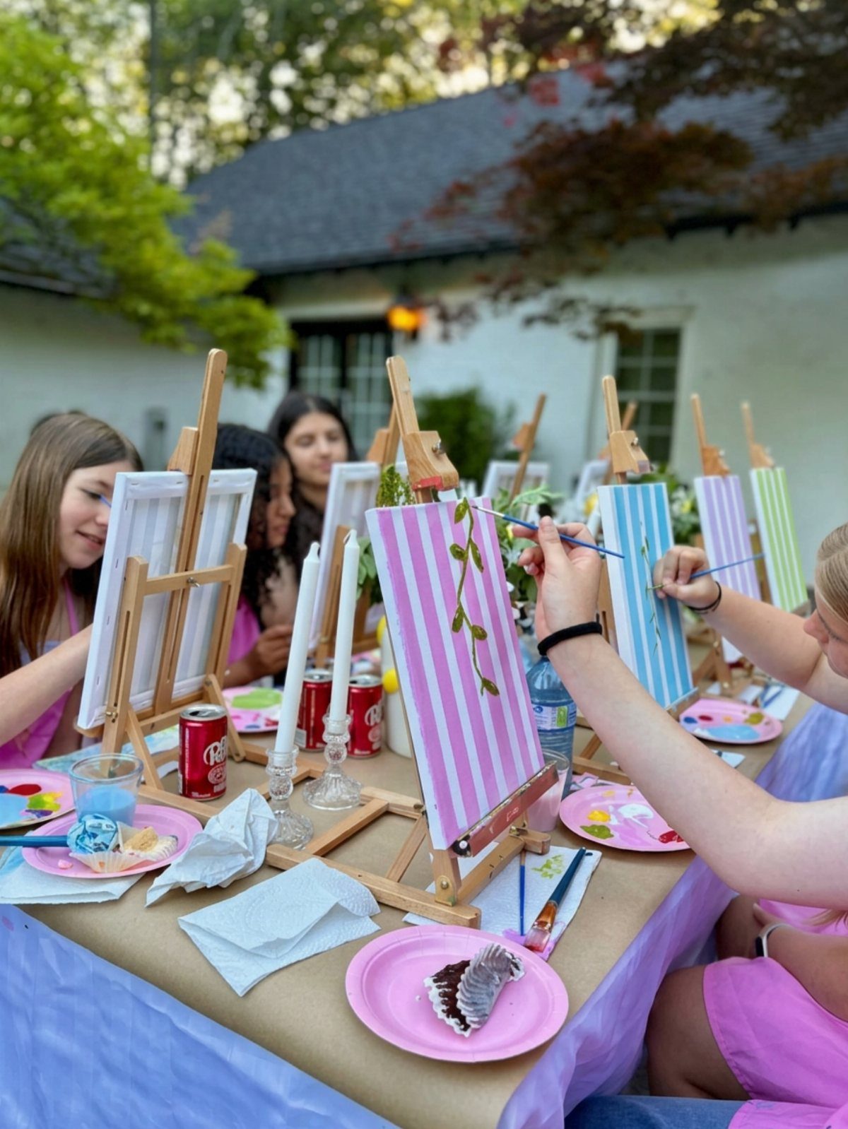 Girls in pink dresses painting on canvases with easels at an outdoor party table