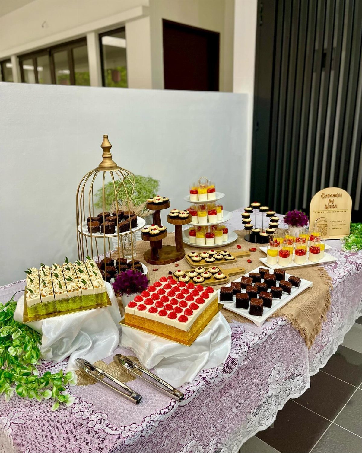 Dessert table display with cakes, brownies, and parfaits on a purple lace tablecloth.
