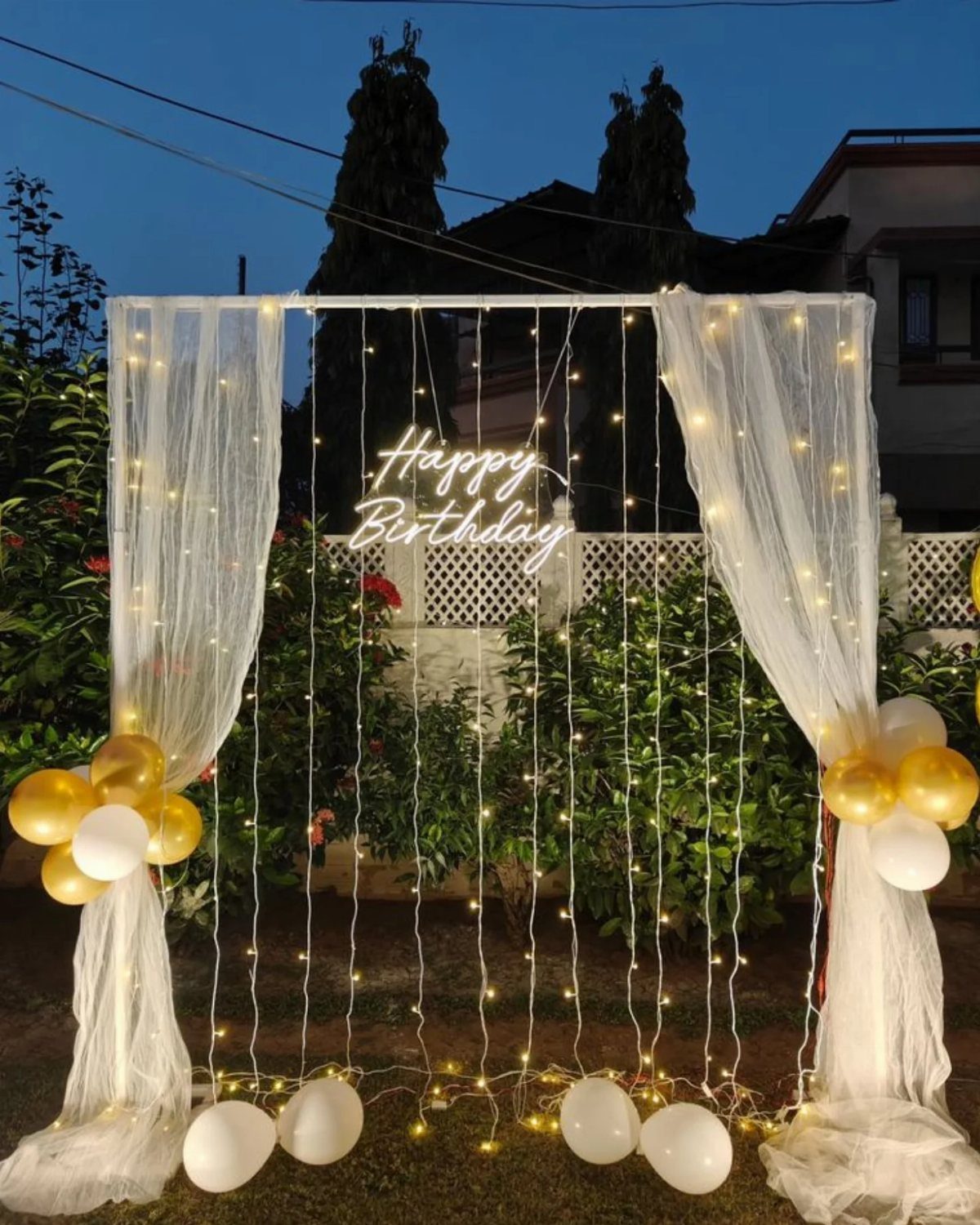 Birthday backdrop with white sheer curtains, fairy lights, gold balloons, and neon sign.