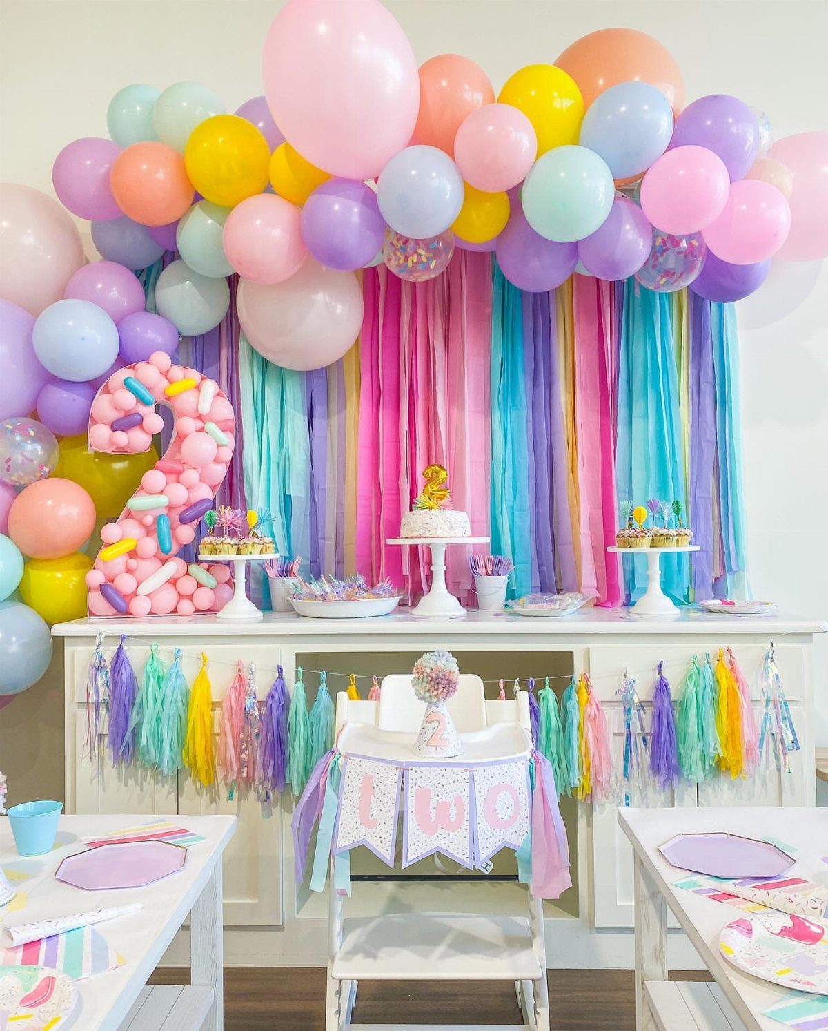 Colorful birthday party setup with pastel balloons, sprinkle-covered number two, and dessert table.