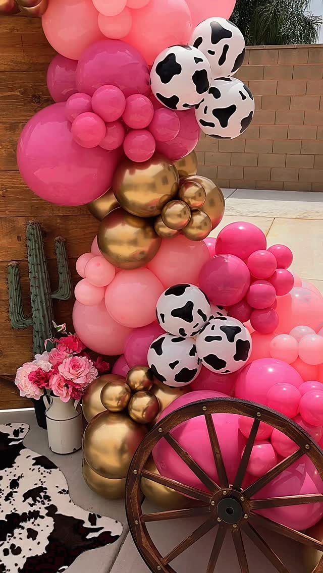 Pink, gold, and cow-print balloon arch with a wooden wagon wheel and flowers.