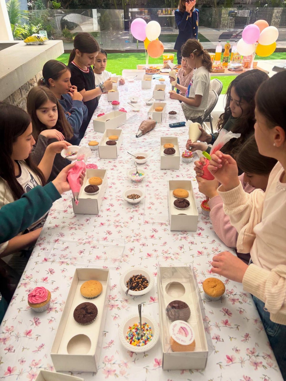 Girls decorating cupcakes with pink frosting and sprinkles at a long outdoor table.
